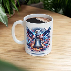 May include: White ceramic coffee mug featuring a patriotic design. The design includes a bald eagle, a red, white, and blue bow, and a Liberty Bell. The mug is filled with dark coffee and has a white handle.