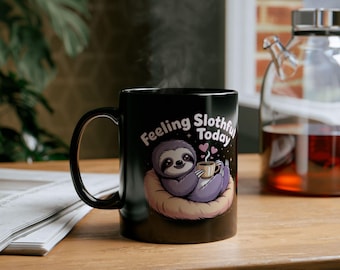 May include: Black ceramic mug with a cartoon sloth design. The sloth, in purple, is holding a coffee cup, with the text "Feeling Slothful Today." The sloth is resting on a brown nest. Steam rises from the mug, suggesting a hot drink.
