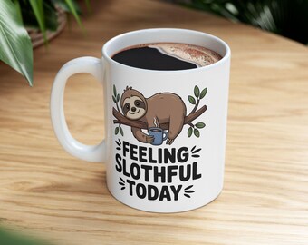 May include: White ceramic coffee mug featuring a brown sloth illustration on a branch, holding a blue mug. The mug is filled with dark coffee and has the text "FEELING SLOTHFUL TODAY" in black. The mug has a white handle.