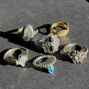 May include: A collection of six rings in various designs. The rings include a gold band, a silver ring with a blue teardrop stone, and a silver ring with a star-shaped stone. Other rings feature black stones and iridescent inlays.