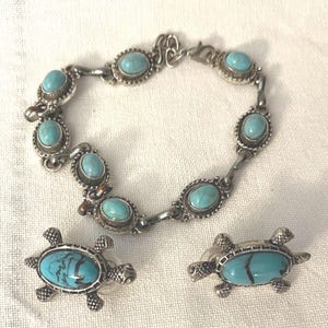 May include: A silver-toned bracelet and matching earrings set. The bracelet features oval turquoise stones set in a silver frame. The earrings are turtle-shaped with turquoise centers. The jewelry is displayed on a white surface.