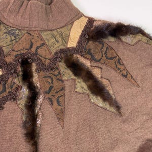 May include: A brown turtleneck sweater with decorative patches of faux snakeskin, fur, and other textured materials. The sweater has a high collar and a unique, artistic design with various shades of brown and tan.