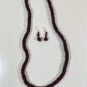 May include: A necklace and matching earrings set. The necklace and earrings are made of small, dark red beads. The necklace is long and hangs in a U-shape. The earrings are small and dangle from silver hooks. The jewelry set is displayed on a white surface.