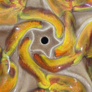 May include: Close-up of a ceramic bundt pan with a star-shaped center and swirling, abstract floral designs. The pan features a color palette of yellow, orange, and brown, with a glossy finish. The center has a hole.