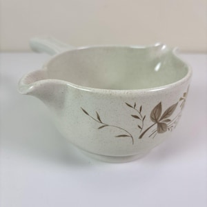 May include: Cream-colored gravy boat with a handle and spout. The ceramic boat features a speckled texture and is decorated with brown leaf and vine designs. A classic serving dish for sauces and gravies.
