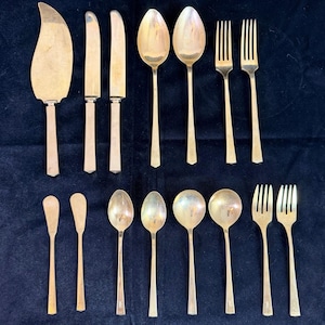 May include: A set of fourteen silver-toned flatware pieces including a cake server, two knives, six spoons of varying sizes, and four forks. Each piece features a smooth, metallic finish with a simple, streamlined handle design.