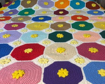 Puede incluir: Una manta de ganchillo hecha a mano con un diseño floral colorido. La manta está compuesta por varias formas hexagonales en tonos de rojo, rosa, morado, amarillo y azul, cada una con un centro de flor amarilla. La manta está sobre una cama.