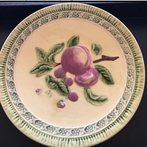 May include: A decorative ceramic plate with a cream-colored center featuring a painted design of purple plums and green leaves. The rim has a detailed border with a green and white floral pattern. The plate is round.