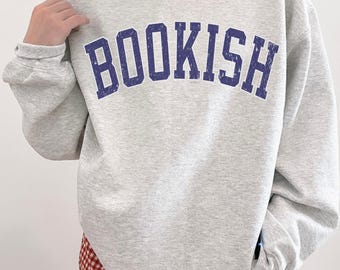 Könnte beinhalten: Ein hellgraues Sweatshirt mit Rundhalsausschnitt und dem Wort BOOKISH, das in einer dunkelblauen, verwaschenen, bogenförmigen Schrift auf der Brust gedruckt ist. Das langärmelige Kleidungsstück hat gerippte Bündchen.