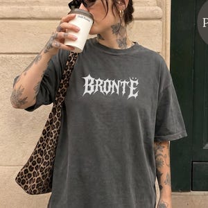 May include: A charcoal grey short-sleeve t-shirt featuring the word BRONTË printed in a white gothic-style font across the chest. The shirt has a relaxed fit and a crew neckline. A leopard print shoulder bag is worn over one side of the garment.