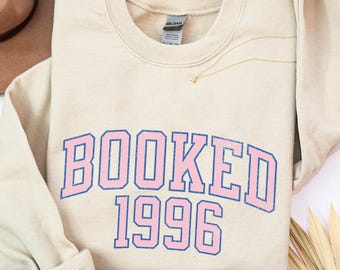 Könnte beinhalten: Ein sandfarbenes Sweatshirt mit Rundhalsausschnitt und dem Text BOOKED 1996 in einer Schriftart im College-Stil. Die Buchstaben sind rosa mit einer blauen Umrandung. Das Kleidungsstück hat lange Ärmel mit gerippten Bündchen und ein Gildan-Etikett.