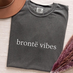 Puede incluir: Una camiseta gris carbón de manga corta con cuello redondo y el texto blanco "brontë vibes" impreso en el pecho. La prenda cuenta con una etiqueta de la marca Comfort Colors en el cuello interior y está hecha de algodón opaco.