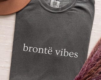 May include: A charcoal gray short-sleeved t-shirt with a crew neckline and the white text "brontë vibes" printed across the chest. The shirt features a Comfort Colors brand label at the inner neck and is made of a solid, opaque cotton fabric.