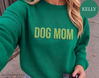 Könnte beinhalten: Ein kellygrüner Rundhals-Pullover mit dem Schriftzug DOG MOM in erhabenen, hellgrünen Blockbuchstaben auf der Brust. Das Kleidungsstück hat lange Ärmel und einen gerippten Ausschnitt sowie ein kreisförmiges Etikett in der Ecke mit der Aufschrift KELLY.