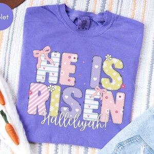 May include: A violet short-sleeved t-shirt featuring the text HE IS RISEN in large, multi-colored, patterned block letters decorated with small flowers, hearts, and pink bows. The word Hallelujah is printed in white cursive script below the text.