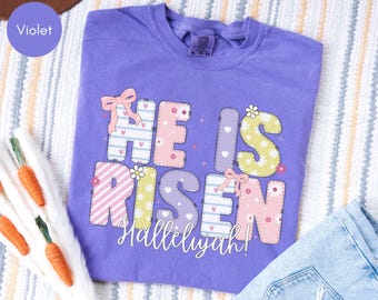 Puede incluir: Una camiseta violeta de manga corta con el texto HE IS RISEN en grandes letras de bloque multicolores y estampadas, decoradas con pequeñas flores, corazones y lazos rosas. La palabra Hallelujah está impresa en cursiva blanca debajo del texto.