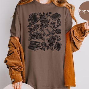 May include: An espresso brown t-shirt featuring a dark brown graphic print of various books and floral rose patterns. The design includes stacked books, open books, and blooming roses with leaves arranged in a central cluster on the front.