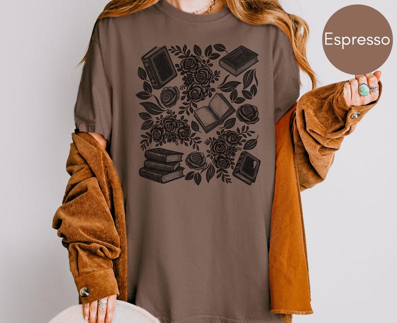 May include: An espresso brown t-shirt featuring a dark brown graphic print of various books and floral rose patterns. The design includes stacked books, open books, and blooming roses with leaves arranged in a central cluster on the front.