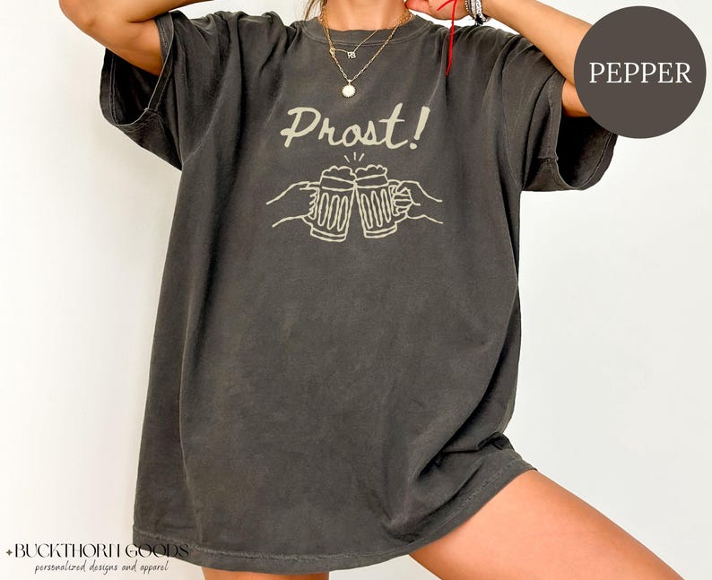 Puede incluir: Una camiseta de manga corta en color gris carb&oacute;n con un estampado gr&aacute;fico claro de dos jarras de cerveza chocando y el texto "Prost!" encima. La prenda presenta un cuello redondo y una silueta holgada y relajada.