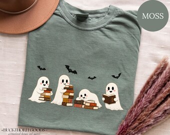 Könnte beinhalten: Ein moosgrünes T-Shirt mit Rundhalsausschnitt und einer Grafik von vier weißen Geistern, die mit Bücherstapeln und offenen Büchern interagieren. Das Design enthält vier kleine schwarze Fledermäuse, die über den Geistern fliegen.
