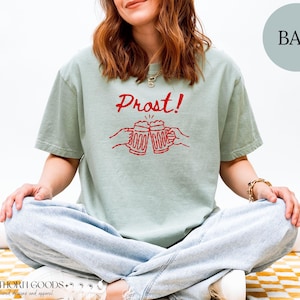 Puede incluir: Una camiseta de manga corta en color verde salvia con un estampado gr&aacute;fico rojo de dos manos brindando con jarras de cerveza y el texto Prost! sobre ellas. La prenda cuenta con cuello redondo y un corte holgado para uso diario.