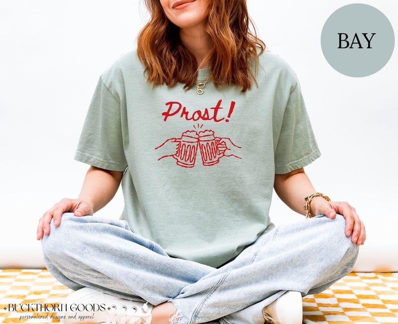 Puede incluir: Una camiseta de manga corta en color verde salvia con un estampado gr&aacute;fico rojo de dos manos brindando con jarras de cerveza y el texto Prost! sobre ellas. La prenda cuenta con cuello redondo y un corte holgado para uso diario.