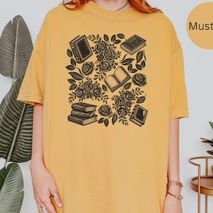May include: A mustard yellow t-shirt featuring a black graphic print of several books and blooming roses. The design includes a stack of three books, two individual closed books, one open book, and various floral elements with leaves scattered throughout.