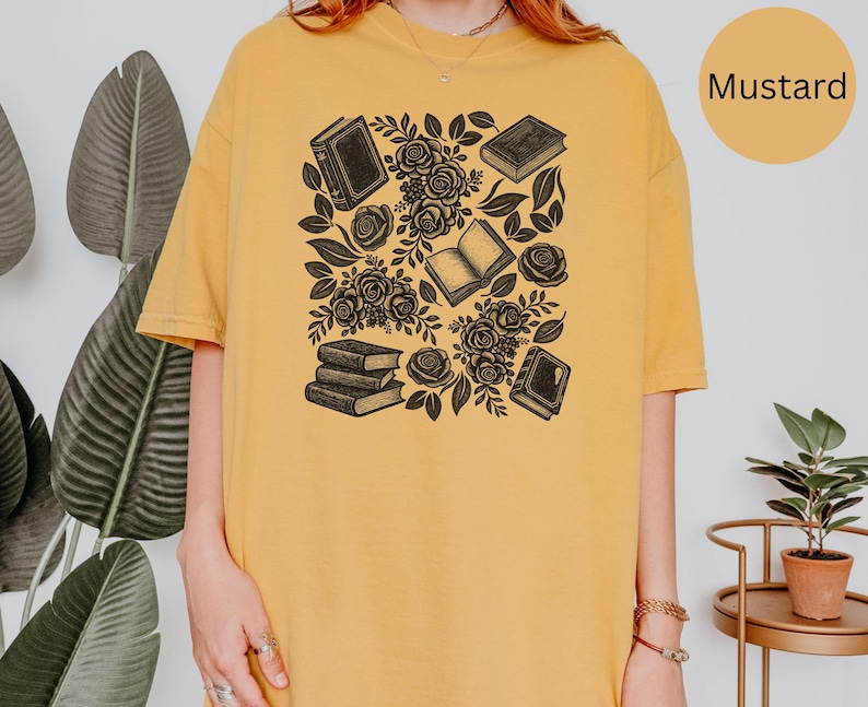 May include: A mustard yellow t-shirt featuring a black graphic print of several books and blooming roses. The design includes a stack of three books, two individual closed books, one open book, and various floral elements with leaves scattered throughout.