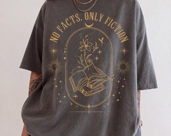 Könnte beinhalten: Ein anthrazitfarbenes T-Shirt mit einem goldenen Grafikdruck eines offenen Buches, aus dessen Seiten eine Blume wächst, umgeben von Sternen und Sonnen-Symbolen. Der Text "NO FACTS, ONLY FICTION" ist über dem ovalen Rahmen gewölbt.