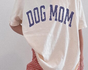 Könnte beinhalten: Ein elfenbeinfarbenes Kurzarm-T-Shirt mit dem Schriftzug DOG MOM in einer abgenutzten marineblauen College-Schrift auf der Brust. Das T-Shirt wird mit einer rot-weiß karierten Shorts kombiniert. Der Text BUCKTHORN GOODS ist sichtbar.