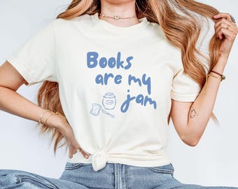 May include: Cream-colored t-shirt with the blue text "Books are my jam" and a small illustration of a jar of jam, a slice of bread, and a knife. The shirt is tied at the waist.