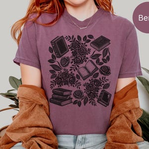 May include: A berry-colored short-sleeve t-shirt featuring a black graphic print of several books and floral rose arrangements. The design includes multiple individual books and a small stack of three books interspersed with detailed rose blossoms.