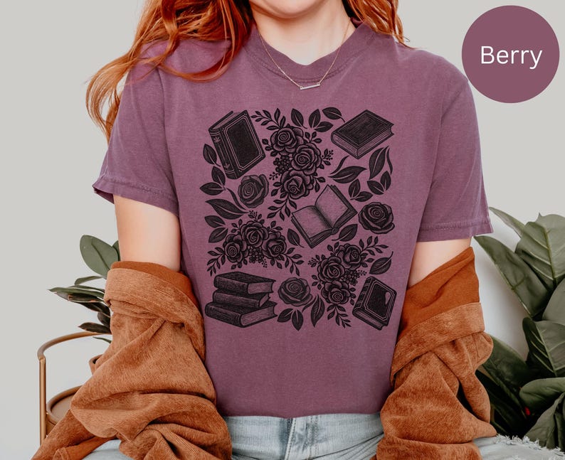 May include: A berry-colored short-sleeve t-shirt featuring a black graphic print of several books and floral rose arrangements. The design includes multiple individual books and a small stack of three books interspersed with detailed rose blossoms.