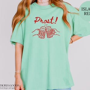 Puede incluir: Una camiseta de manga corta en color verde menta claro con un estampado gr&aacute;fico rojo de dos manos brindando con jarras de cerveza y el texto Prost! centrado encima. El dise&ntilde;o est&aacute; impreso en tinta roja sobre la parte frontal.