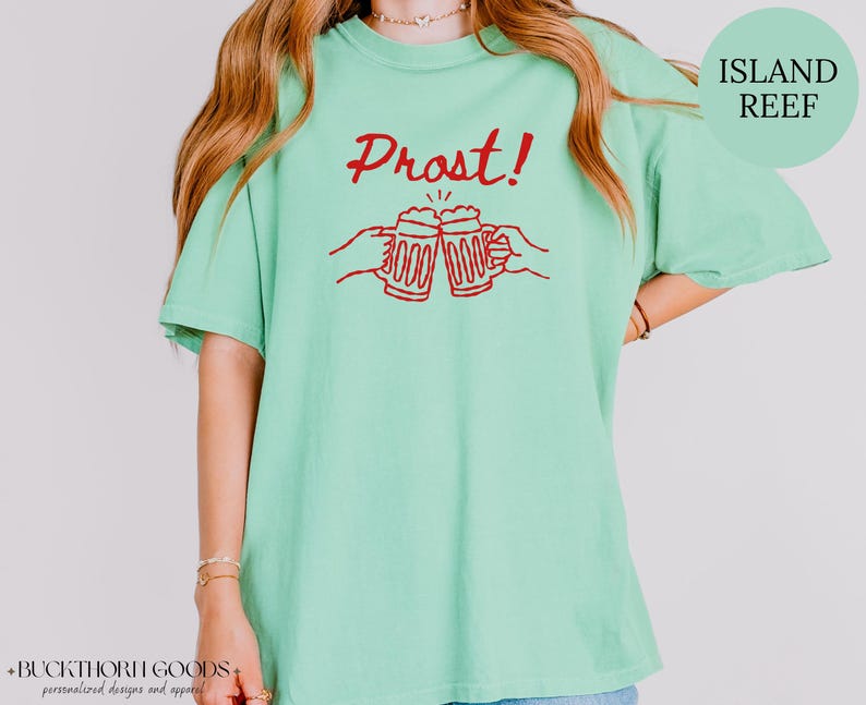 Puede incluir: Una camiseta de manga corta en color verde menta claro con un estampado gr&aacute;fico rojo de dos manos brindando con jarras de cerveza y el texto Prost! centrado encima. El dise&ntilde;o est&aacute; impreso en tinta roja sobre la parte frontal.
