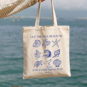 May include: A beige tote bag with blue illustrations of seashells, coral, and a whale tail. The text "LET THE SEA RESOUND AND EVERYTHING IN IT" is printed in blue. The bag is held up against a blurred ocean background.