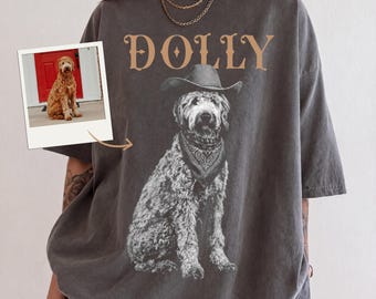 Könnte beinhalten: Ein anthrazitfarbenes Kurzarm-T-Shirt mit einer Grafik eines Hundes, der einen Cowboyhut und ein Halstuch trägt. Der Text DOLLY ist in Beige über dem Hund gedruckt. Ein kleines Foto zeigt denselben Hund vor einer roten Tür sitzend.