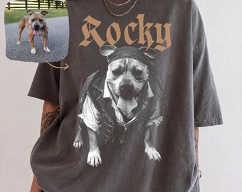 Könnte beinhalten: Ein anthrazitfarbenes Kurzarm-T-Shirt mit einer großen Grafik eines Hundes, der ein Bandana und eine Weste trägt. Der Text Rocky ist in einer beigen gotischen Schrift über dem Hund gedruckt. Ein kleines Bild zeigt den Hund auf einem Weg.