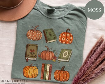 Könnte beinhalten: Ein moosgrünes T-Shirt mit einem Grafikdruck aus verschiedenen orangefarbenen Kürbissen und Büchern im Vintage-Stil. Das Design zeigt Kürbisse mit Punkt- und Blumenmustern sowie geschlossene Bücher mit botanischen Illustrationen und verzierten Buchrücken.