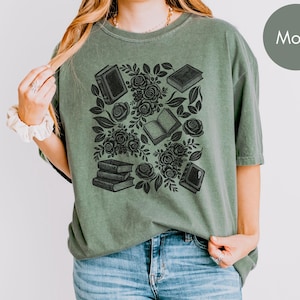 May include: A moss green short-sleeve t-shirt featuring a black graphic print of stacked books, open books, and blooming roses with leaves. The shirt has a crew neckline and a relaxed fit, showcasing a detailed botanical and literary illustration.