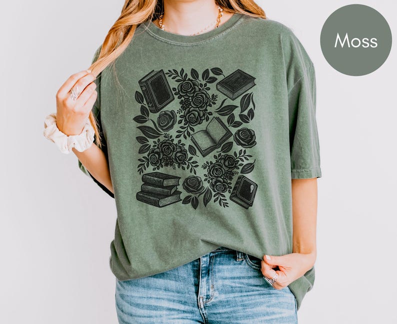 May include: A moss green short-sleeve t-shirt featuring a black graphic print of stacked books, open books, and blooming roses with leaves. The shirt has a crew neckline and a relaxed fit, showcasing a detailed botanical and literary illustration.