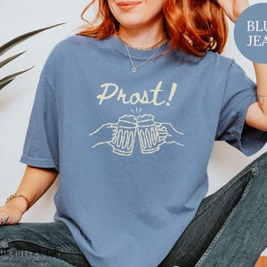 Puede incluir: Una camiseta de manga corta color azul jean con un estampado gr&aacute;fico en color crema de dos manos brindando con jarras de cerveza y el texto "Prost!" encima. La prenda cuenta con cuello redondo y un corte holgado de manga corta.