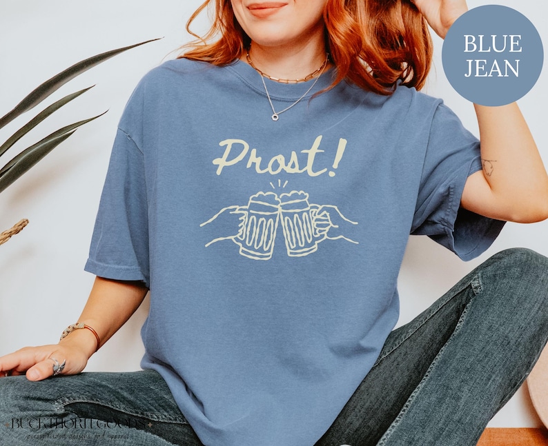 Puede incluir: Una camiseta de manga corta color azul jean con un estampado gr&aacute;fico en color crema de dos manos brindando con jarras de cerveza y el texto "Prost!" encima. La prenda cuenta con cuello redondo y un corte holgado de manga corta.