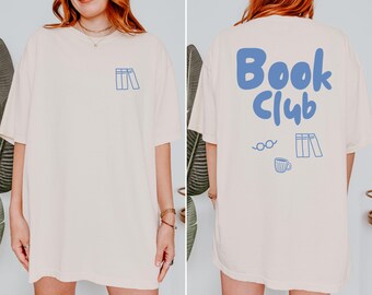 May include: An oversized cream-colored t-shirt featuring a small blue line-art graphic of books on the front. The back displays the text "Book Club" in large blue letters above line-art illustrations of eyeglasses, a coffee mug, and books.