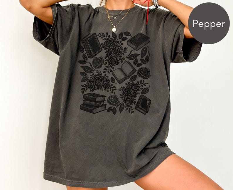 May include: A charcoal gray short-sleeve t-shirt featuring a black graphic print of stacked books, open books, and blooming roses. The shirt has a crew neckline and a relaxed fit. The word Pepper is printed in white on a dark gray circular background.