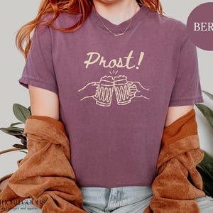 Puede incluir: Una camiseta de manga corta color baya con un gr&aacute;fico en color crema de dos jarras de cerveza chocando entre s&iacute;, con el texto Prost! impreso encima. La camiseta tiene cuello redondo y un corte relajado con un dise&ntilde;o gr&aacute;fico sencillo.