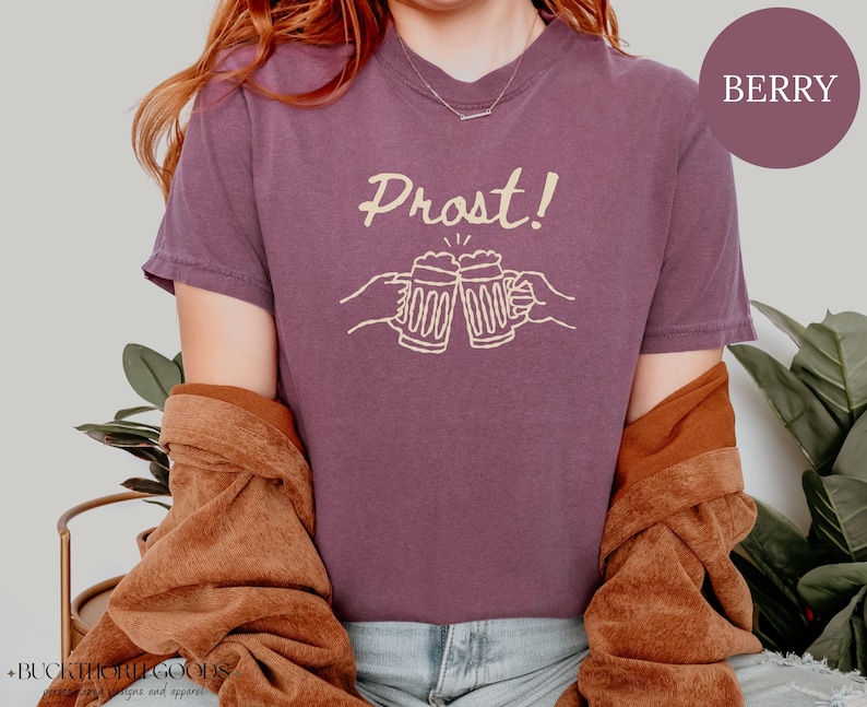 Puede incluir: Una camiseta de manga corta color baya con un gr&aacute;fico en color crema de dos jarras de cerveza chocando entre s&iacute;, con el texto Prost! impreso encima. La camiseta tiene cuello redondo y un corte relajado con un dise&ntilde;o gr&aacute;fico sencillo.