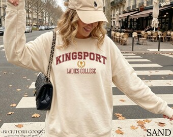 Könnte beinhalten: Ein sandfarbenes Sweatshirt mit Rundhalsausschnitt, das den Schriftzug KINGSPORT LADIES COLLEGE in kastanienbraunen Buchstaben und ein goldenes Lorbeerkranz-Emblem zeigt. Dazu eine beige Baseballkappe und eine schwarze gesteppte Umhängetasche.