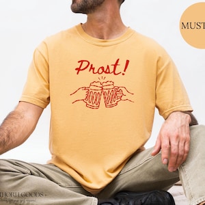 Puede incluir: Una camiseta de manga corta color amarillo mostaza con un gr&aacute;fico rojo de dos jarras de cerveza chocando y el texto "Prost!" impreso encima. La camiseta tiene cuello redondo y un corte relajado con un dise&ntilde;o de l&iacute;neas sencillo.