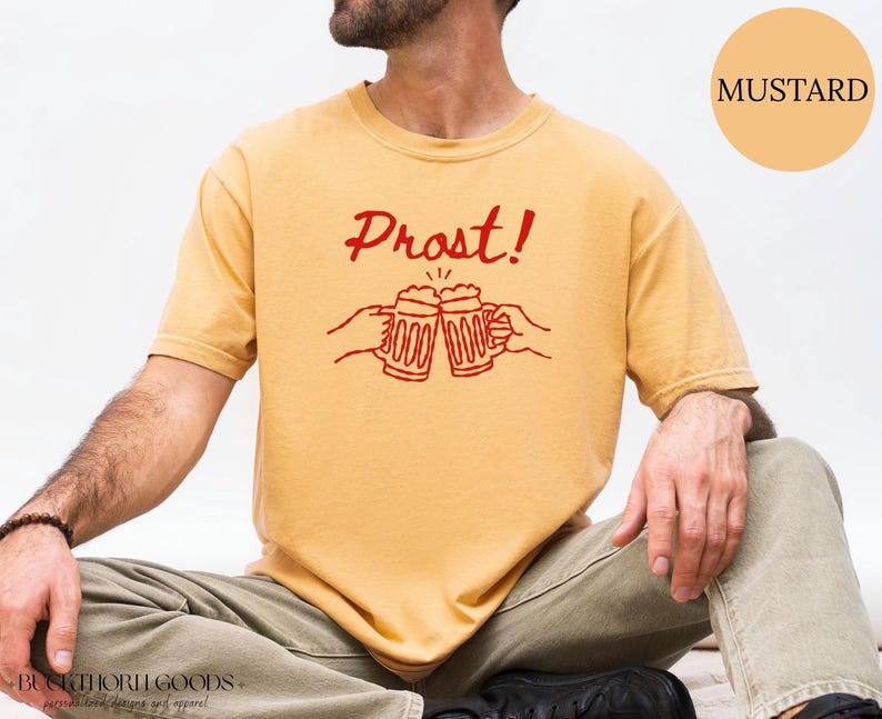 Puede incluir: Una camiseta de manga corta color amarillo mostaza con un gr&aacute;fico rojo de dos jarras de cerveza chocando y el texto "Prost!" impreso encima. La camiseta tiene cuello redondo y un corte relajado con un dise&ntilde;o de l&iacute;neas sencillo.